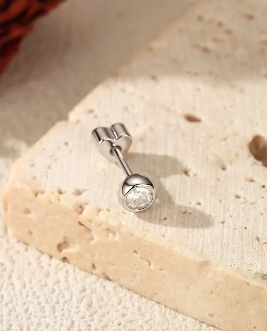 Puces d’Oreilles Moissanite Serti Clos en Argent 925 - posée sur une surface claire texturée près d’une dalle de travertin