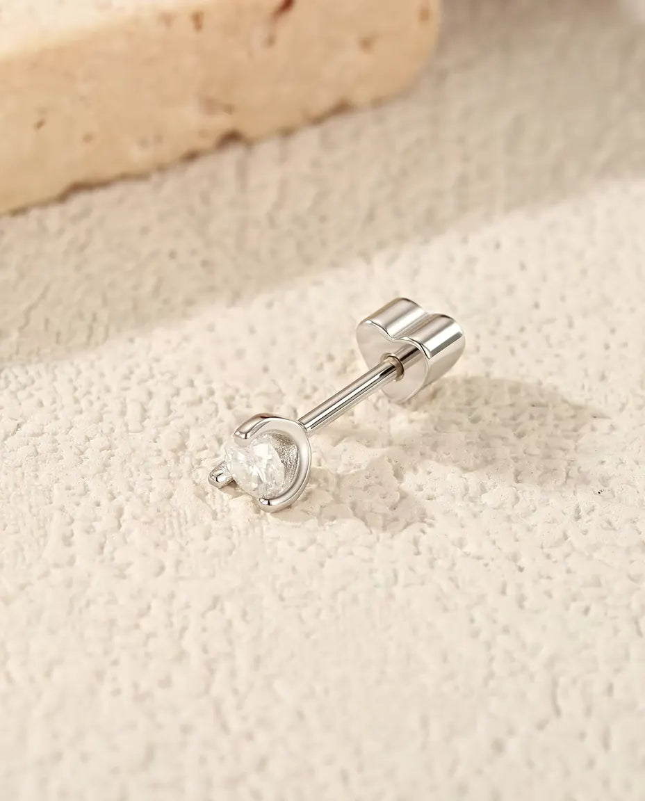 Puces d’Oreilles Moissanite Ronde à Arc Latéral en Argent 925 - posée sur le côté sur une surface claire texturée, fermoir en forme de cœur sur la tige