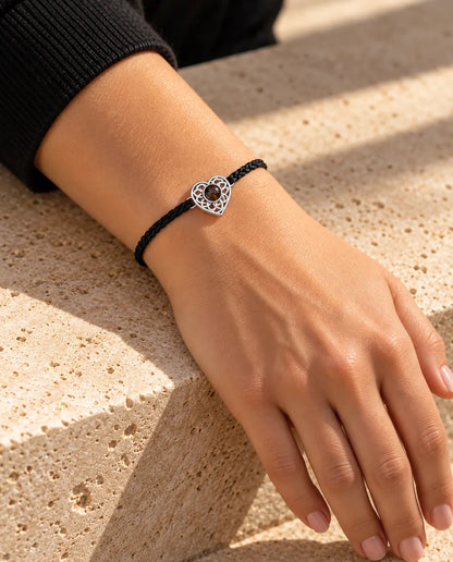 Femme portant le bracelet cordon fin tressé noir avec cœur ajouré argenté au poignet, sous une lumière chaude