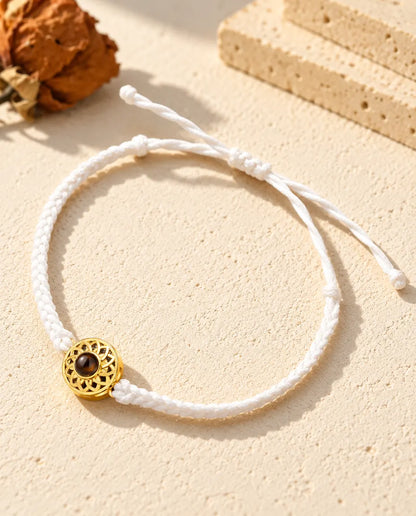 Bracelet cordon fin tressé blanc avec médaillon rond doré et nœud coulissant sur plâtre clair