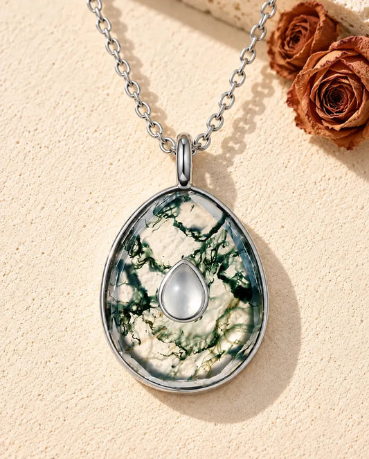 Collier Agate Mousse Pendentif Œuf en Argent 925 - posé sur une surface claire texturée près d’un bloc de travertin