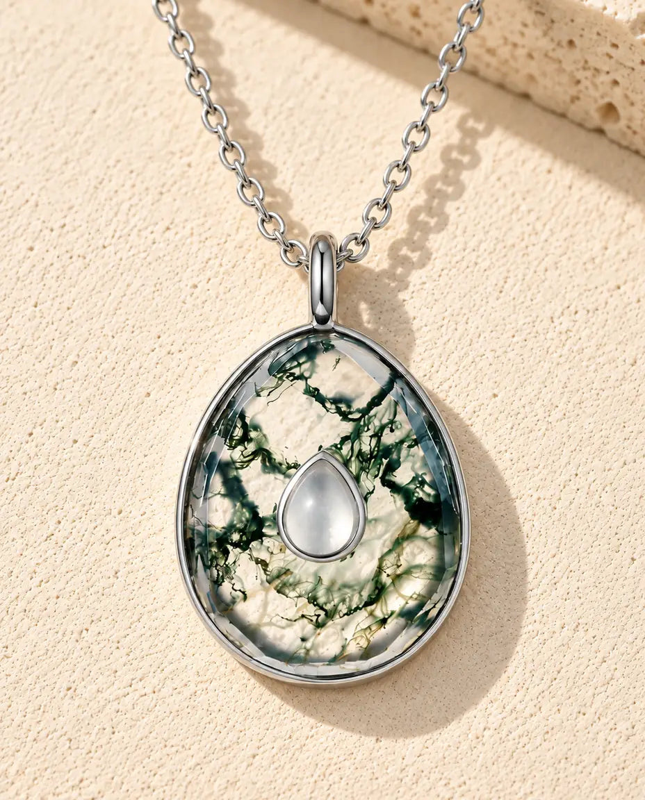 Collier Agate Mousse Pendentif Œuf en Argent 925 - posé sur une surface claire texturée près d’un bloc de travertin