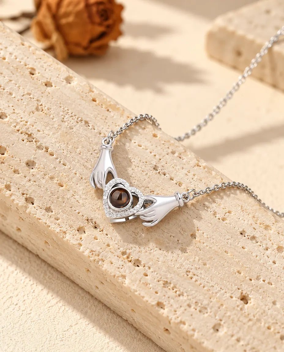 Collier photo projection personnalisé argenté, deux petites mains tenant un cœur pavé de zircons, posé contre du travertin