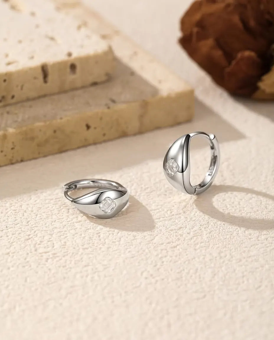 Boucles d’Oreilles Créoles Huggie Moissanite Taille Asscher en Argent 925 - la paire sur une surface claire texturée, l’une à plat et l’autre debout