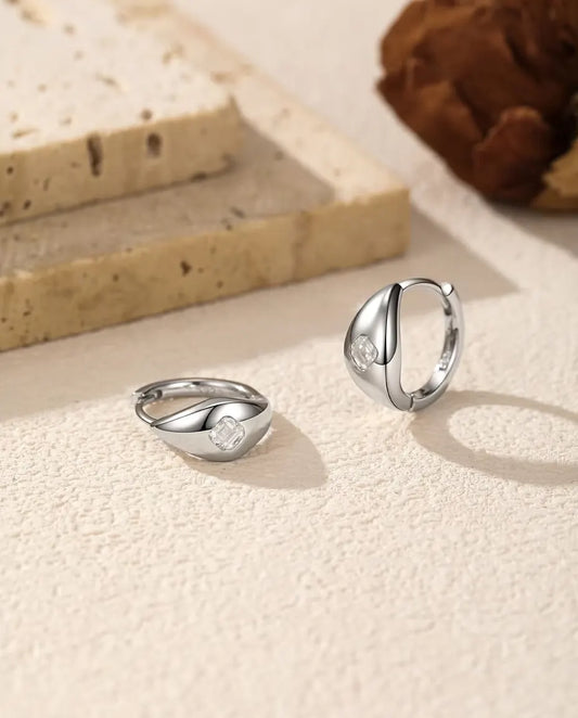 Boucles d’Oreilles Créoles Huggie Moissanite Taille Asscher en Argent 925 - la paire sur une surface claire texturée, l’une à plat et l’autre debout