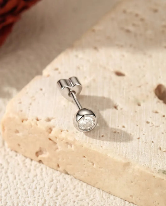 Puces d’Oreilles Moissanite Serti Clos en Argent 925 - posée sur une surface claire texturée près d’une dalle de travertin