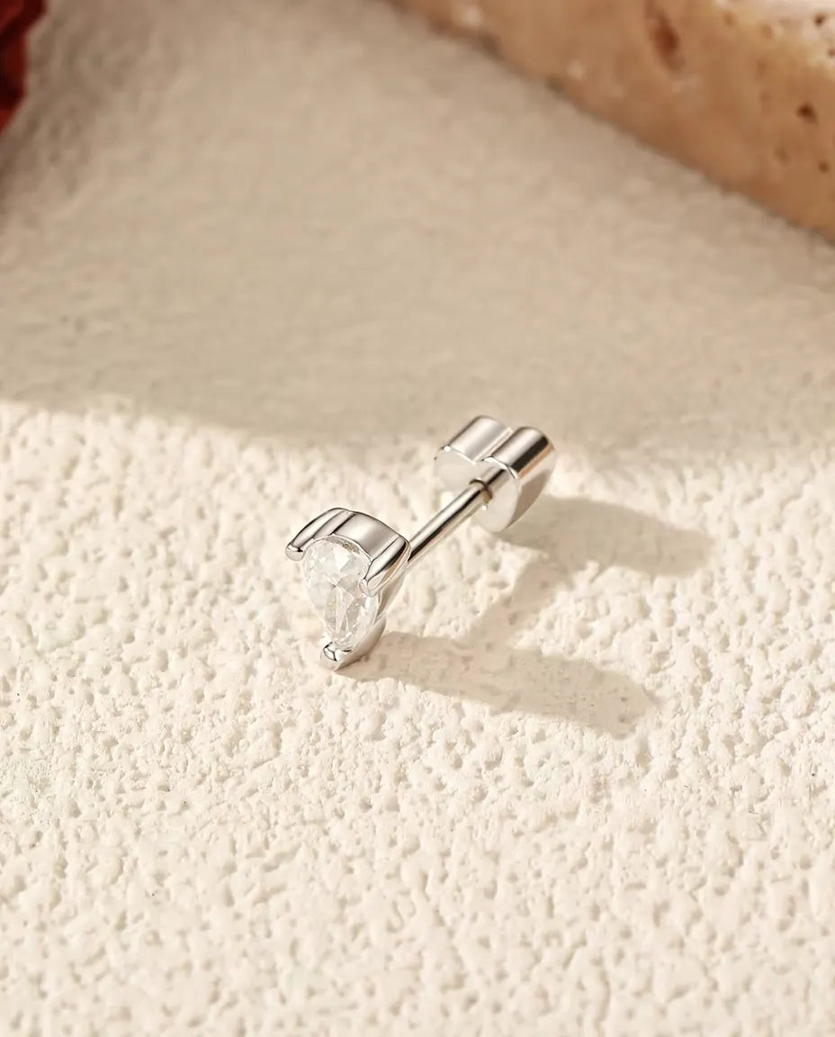Puces d’Oreilles Moissanite Poire à Deux Griffes en Argent 925 - posée sur le côté sur une surface claire texturée, fermoir en forme de cœur sur la tige